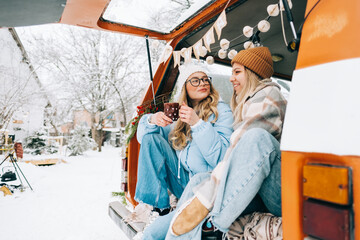 Two cheerful women friends sitting in a van in winter camp and having fun, enjoying holiday.