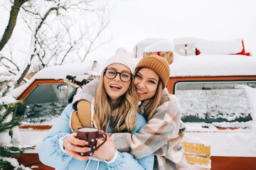 Portrait of two cheerful women friends heaving fun standing outdoor near van, enjoying winter time.