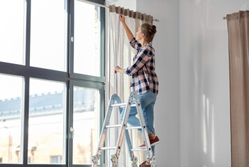 Obraz na płótnie Canvas home improvement, decoration and renovation concept - happy smiling woman on ladder hanging curtains