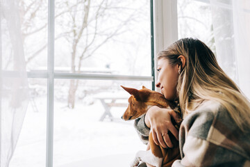 Young caucasian happy woman sitting near window with her dog in winter holidays, looking outside.