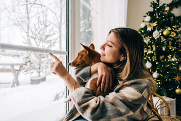 Young caucasian happy woman sitting near window with her dog in winter holidays, looking outside.