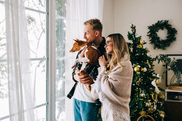 Young couple standing near big window with his dog, enjoying winter holidays.