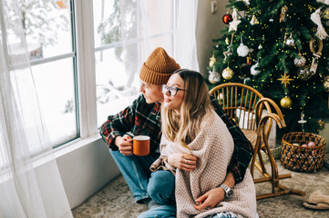 Young couple sitting on a floor near big window and looking outside, enjoying winter holidays.