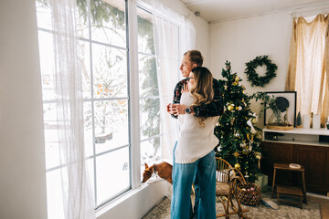 Young couple standing near big window and looking outside, enjoying winter holidays.