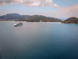 Black and white liner crosses a beautiful blue bay, coastal Thai town on the background; drone shot, side view.