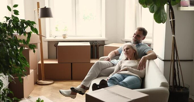 Mature couple talking relaxing on relocation day to new own home seated lying on cozy sofa in living room near boxes with stuff and house plants. Moving, repairs, bank loan for older clients concept
