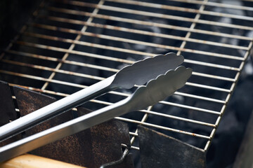 Stainless steel grill tongs on a dirty grill rust. Wooden handle blurred in the foreground. - obrazy, fototapety, plakaty