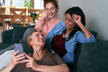 Three best caucasian friends smiling while browsing smart phone   on the sofa