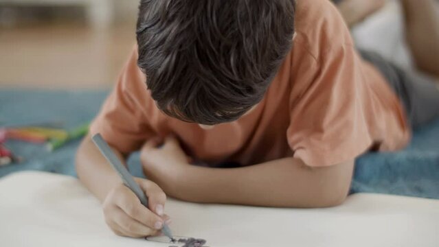 Focused little boy lying on floor at home and painting picture. Close-up shot of concentrated Caucasian kid drawing with felt pen in drawing book, studying alone. Education, hobby concept