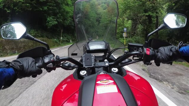 POV Biker rides on a motorbike by forested mountain road in Italy. Motorcyclist on motorcycle goes between mountain green trees by landscape highway. Steering wheel view. First-person view. Moto trip