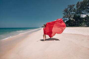 Gorgeous girl in the sunny exotic beach by the ocean. The young woman wears amazing red dress...