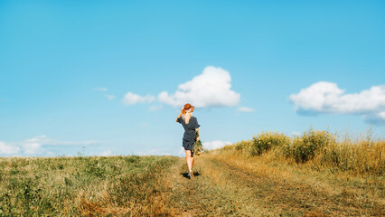 Back view of slim elegant young woman in summer dress and straw hat running on field on horizon on sunny day, outdoors. Beauty caucasian girl, romantic lifestyle