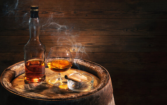Glass of whiskey or cognac, bottle and smoking cigar on vintage wooden barrel