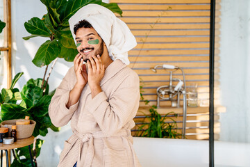 Beauty, spa and leisure concept. Funny asian man with green eyes patches in bathrobe and bath towel over head sitting on bathtub in cozy bathroom.