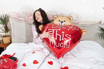 A beautiful woman in pink pajamas with a heart-shaped balloon lies on bed at home