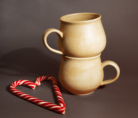 Two ceramic mugs of light yellow color are placed on top of each other, next to candy sticks are folded in the shape of a heart. Handmade ceramics concept, Valentine's Day.