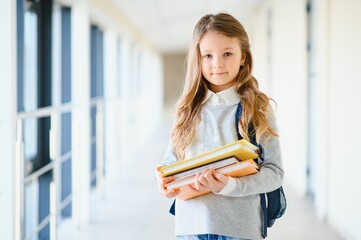 Front view of little beautiful school girl among corridor at school, holding notes at hands. Funny and happy girl smiling at camera, resting after lessons on primary school
