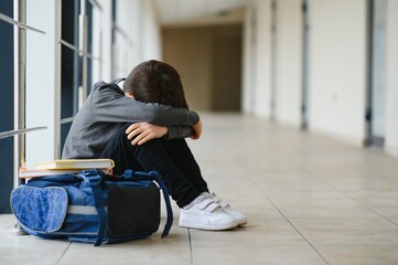 Little boy sitting alone on floor after suffering an act of bullying while children run in the background. Sad young schoolboy sitting on corridor with hands on knees and head between his legs.