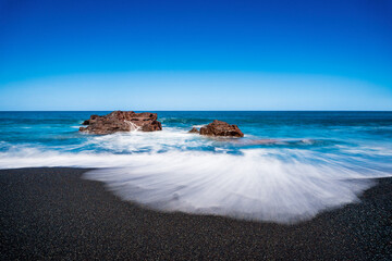 Meerblick mit Felsen am schwarzen Sandstrand mit blauen Himmel und Sonnenschein