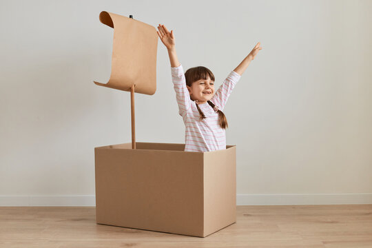 Happy exited Caucasian little dark haired female child making ship from carton box playing alone at home against white wall, raised arms with clenched fists.