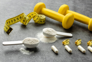 Sports supplements in measuring spoons and capsules close-up next to dumbbells and a measuring tape.