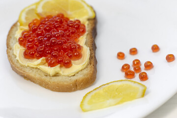 A piece of sliced loaf with butter, red caviar and lemon on a white plate. Bright and juicy photo close up of bread and butter sandwich with red salmon caviar and lemon