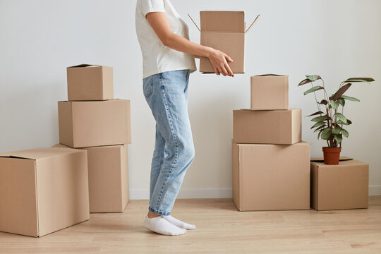 Indoor shot of faceless unknown anonymous woman wearing white t shirt and jeans, holding cardboard box with her belongings during relocation to a new flat.