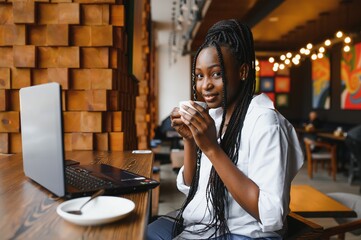 Side view of black lady enjoying morning coffee and checking emails on laptop, cafe interior, empty space
