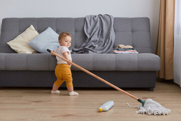 Indoor shot of infant toddler baby wearing yellow pants standing near gray sofa with mop, little charming girl helping mother doing household chores, try to wash floor.