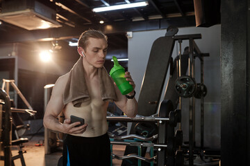 Fototapeta na wymiar Fit young man with towel on his shoulder drinking water after intense training in gym