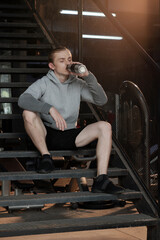 Young man in shorts and hoodie sitting on steps and drinking water after working out in gym