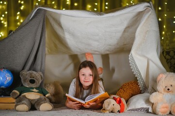 Obraz na płótnie Canvas happy child girl laughing and reading book in dark in a tent at home