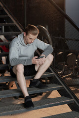 Fototapeta na wymiar Serious sportsman sitting on gym steps and checking sport results on smartphone application after training