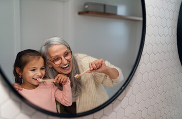 Grandmother with granddaughter standing indoors in bathroom, brusing teeth and looking at mirror.