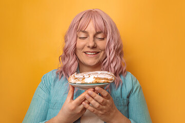 Portrait of beautiful happy woman holding in hands eclair cake isolated over pink wall background.