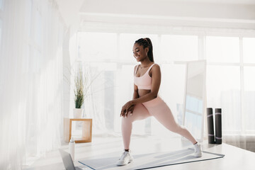 Millennial black woman making lunges near laptop, working out to online sports tutorial at home, free space
