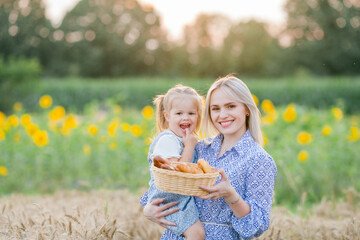 Adorable mother and daughter in romantic dresses eat fresh pastries in a wheat field. Summer family portrait. Eco.