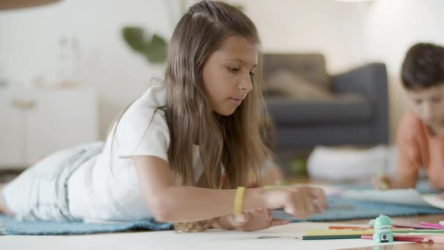 Cute girl lying on floor, drawing picture with colored pencils. Medium shot of focused kid developing creative skills, doing homework, devoting time to hobby. Learning, education concept
