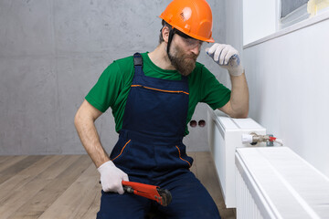 A male plumber installs a radiator in the heating system of an apartment. Guy in overalls and a gas wrench.