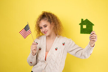 A young girl holds a model of a green house in her hands and an American flag. The concept of buying an eco house.