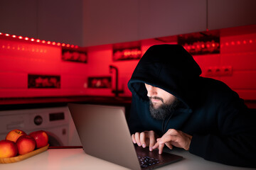 a hacker in a black hoodie with a hood on his head is sitting at a laptop bent over hacking the...
