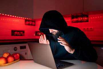 a hacker in a black hoodie with a hood on his head is sitting at a laptop with his palms spread in...