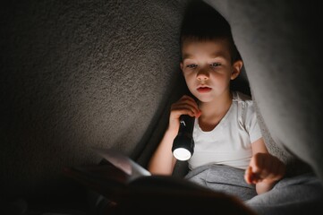Boy with flashlight reading book under blanket at home