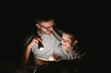 Father and son with flashlight reading book under blanket at home