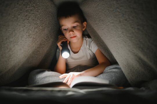 Reading book and using flashlight. Young boy in casual clothes lying down near tent at evening time.