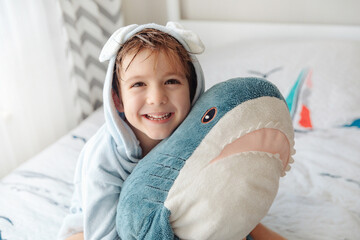 Happy laughing kid boy in soft blue bathrobe after bath play with shark toy on white bed in sunny bedroom. Wash and children's hygiene concept.