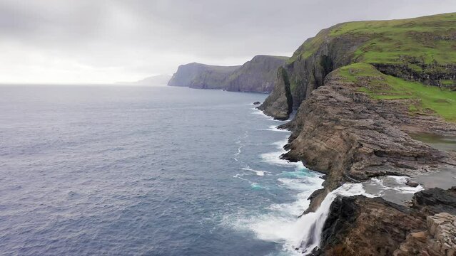 Aerial view of rock cliff in Faroe Islands. Blue sea waves, rough sea on cloudy journey. slow motion