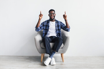 Excited young black man having idea and pointing two fingers up at empty space, sitting in armchair, mockup