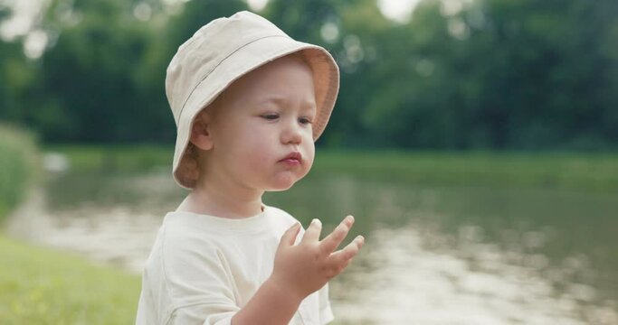 A close-up on a face of a small child wearing a hat, eating fruit, the boy is sitting on the grass near water, lake, a caring father wipes son's face dirty with food