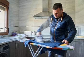 Young man ironing clothes on ironing board in kitchen room